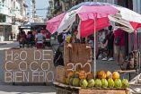 Vendedor ofrece agua de coco bajo sombrilla en una calle de La Habana, con carteles artesanales y peatones alrededor en contexto de crisis económica en Cuba.