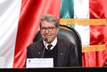 Ricardo Monreal Ávila, coordinador de Morena en el Senado, sonríe mientras habla frente a micrófono con bandera de México de fondo.