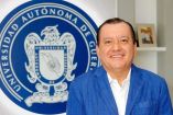 Javier Saldaña con saco azul sonríe frente al emblema oficial de la Universidad Autónoma de Guerrero en una fotografía institucional en interior.