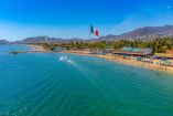 Vista aérea de la playa de Acapulco con visitantes en la franja costera, bandera monumental de México y zona hotelera junto al mar.