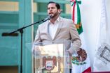 Ismael Burgueño Ruiz habla en un podio con escudo de Tijuana durante un acto oficial, con bandera de México al fondo y micrófono