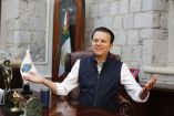 Esteban Villegas Villarreal sentado en su oficina en Durango, con bandera de México al fondo y escritorio con objetos institucionales