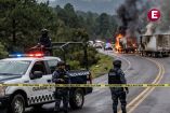 Una patrulla de la Guardia Nacional y agentes de la policía establecen un cordón de seguridad con cinta amarilla en una carretera de Michoacán. En el fondo, varios camiones de carga están incendiándose, emitiendo gruesas columnas de humo negro en un entorno boscoso y montañoso. Imagen representativa de narcobloqueos y violencia en la región.