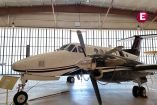 Vista exterior del avión Beechcraft blanco con franjas negras del Mayo Zambada exhibido en un hangar, con gruesas cadenas en la hélice derecha.