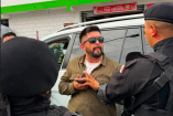 Hombre con gafas de sol conversa con policías junto a una camioneta estacionada frente a un establecimiento comercial.