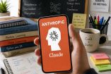 Teléfono móvil muestra el logotipo de Anthropic y Claude sobre un escritorio con libros y apuntes relacionados con inteligencia artificial.