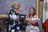 Angela Davis recibe un reconocimiento junto a una funcionaria durante un acto oficial en la Ciudad de México, frente a la bandera nacional.