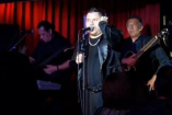 Alejandro Zúñiga interpreta sus canciones durante una presentación en vivo, acompañado por músicos, ante el público y con iluminación escénica.