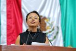 Diputada local con lentes amarillos habla desde la tribuna legislativa, frente a una bandera de México durante una sesión pública.