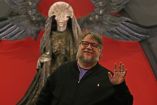 Guillermo del Toro sonríe y saluda frente a una figura monstruosa con alas negras y fondo rojo.