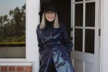 Diane Keaton con sombrero negro, lentes y abrigo azul brillante, sonriendo frente a una puerta blanca.