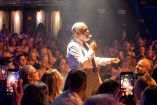 Francisco Céspedes canta con micrófono entre el público, vestido con camisa blanca y corbata negra, mientras asistentes lo graban.