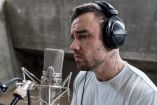 Liam Payne con audífonos negros y camiseta gris canta frente a un micrófono de estudio, con tatuajes visibles en el cuello.