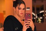 Selena Gomez se toma una selfie frente al espejo con un celular morado, bata negra y cabello recogido.