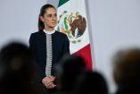 La presidenta Claudia Sheinbaum escucha atentamente durante su conferencia de prensa matutina, de pie frente a la bandera de México.