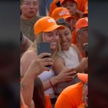 Dos mujeres sonrientes, una tomando una selfie, en un evento de campaña, rodeadas de seguidores con gorras naranjas del partido Movimiento Ciudadano, simbolizando la participación ciudadana y el apoyo a los emprendedores locales en Monterrey.