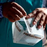 Primer plano de las manos de un representante indígena wixárika manipulando una urna electoral con la palabra "Presidencia" durante una capacitación del INE en Tuxpan de Bolaños, Jalisco, México, antes de las elecciones presidenciales.