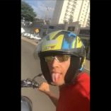 Motociclista joven tomando una selfie con casco y gafas de sol mientras conduce por una vía urbana en São Paulo, Brasil.