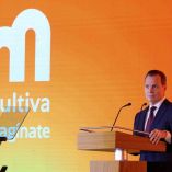 Olegario Vázquez Aldir, presidente de Grupo Vazol, en el escenario durante la presentación oficial de la nueva identidad gráfica de Grupo MultiVA, con un fondo naranja que muestra el nuevo logotipo e identidad visual de la marca.