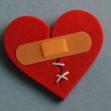 Ilustración de un corazón roto de fieltro rojo, parcialmente cosido con hilo blanco y cubierto por una curita color piel. La imagen representa el proceso de sanar un corazón roto, en referencia al Día Internacional del Mal de Amores, sobre un fondo azul que acentúa el contraste emocional de la escena