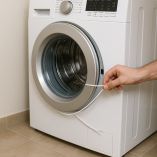 Mano masculina usando un cordón blanco para intentar desbloquear la puerta de una lavadora de carga frontal. El cordón rodea el aro de la puerta mientras se aplica presión hacia el cierre, en un entorno doméstico con piso de cerámica.