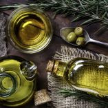 Vista superior de frascos con aceite de oliva virgen extra, una cuchara con aceitunas verdes y ramas de romero sobre una mesa de madera rústica. La imagen representa ingredientes clave de la dieta mediterránea y hace alusión a los beneficios nutricionales y metabólicos del aceite de oliva según estudios del CSIC.