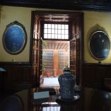 Vista interior de la sala principal del Hospital de Jesús en el Centro Histórico de la Ciudad de México, con retratos antiguos de Hernán Cortés, mobiliario virreinal, jarrón de talavera sobre una mesa circular y acceso a un patio central con puertas de madera y luz natural.