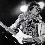 Fotografía en blanco y negro de Jimi Hendrix cantando y tocando la guitarra eléctrica durante un concierto en los años sesenta. La imagen captura su energía en escena, con vestimenta psicodélica y luces de fondo. Es parte del contexto de la demanda legal por derechos de intérpretes en la era del streaming.