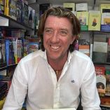 Colate Vallejo-Nágera sonríe durante su visita a la Feria del Libro, posando ante un expositor repleto de libros en un entorno cultural y literario.