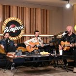 Artistas interpretan canciones en vivo en un set íntimo de SONGBOOK, rodeados de instrumentos acústicos y micrófonos, durante una sesión que prioriza la emoción, la interpretación real y la conexión musical.