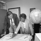 Gladys West, matemática estadounidense y pionera del GPS, revisa mapas y documentos técnicos junto a un colega en una oficina, con globos terráqueos y material cartográfico al fondo.