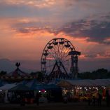 Vista nocturna de la Feria Santa Inés en Zacatelco, Tlaxcala, con juegos mecánicos iluminados y una rueda de la fortuna en primer plano, mientras el cielo al atardecer tiñe de rojo y naranja el horizonte volcánico.