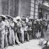 Fotografía histórica en blanco y negro de un grupo numeroso de trabajadores rurales formados en fila frente a un edificio de piedra, mientras una mujer organiza o reparte atención, escena representativa del contexto social y laboral en México.