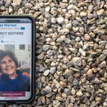 Teléfono celular mostrando la ficha de Nancy Guthrie en la página “Most Wanted” del FBI, apoyado sobre grava en Tucson, Arizona, en medio de la búsqueda por la desaparición de la madre de la periodista Savannah Guthrie.