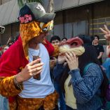Joven con gorro de cabeza de perro y chamarra roja interactúa con joven que porta máscara de dinosaurio durante la reunión therian en la Explanada de Rectoría de la UANL, ante la mirada de otros estudiantes.