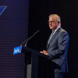 Anthony Albanese pronuncia un discurso en el Future Victoria Summit en Melbourne, de pie tras un atril con micrófono y teleprompter, en un escenario con iluminación tenue y fondo oscuro.