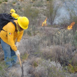 Brigadista con casco y uniforme amarillo trabaja con herramienta manual para contener un incendio forestal en zona de matorral en Múzquiz, Coahuila, mientras al fondo se observan llamas activas entre la vegetación seca.