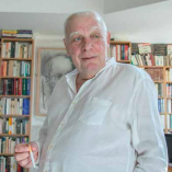 António Lobo Antunes de pie en su casa en Lisboa frente a estanterías llenas de libros, sosteniendo un cigarrillo mientras observa hacia un costado.