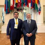 Pedro Haces Barba posa junto a representante internacional en sede de la OEA en Washington, bajo banderas de países miembros durante agenda sobre democracia.
