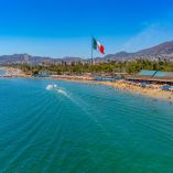 Vista aérea de la playa de Acapulco con visitantes en la franja costera, bandera monumental de México y zona hotelera junto al mar.