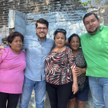 Irugami Perea Cruz posa junto a cuatro habitantes frente a una vivienda de ladrillo en una colonia de Acapulco durante una visita comunitaria.