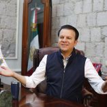 Esteban Villegas Villarreal sentado en su oficina en Durango, con bandera de México al fondo y escritorio con objetos institucionales
