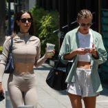 Dos mujeres jóvenes caminan por una calle urbana soleada con ropa ligera, bebidas frías y teléfono móvil en mano.