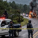Una patrulla de la Guardia Nacional y agentes de la policía establecen un cordón de seguridad con cinta amarilla en una carretera de Michoacán. En el fondo, varios camiones de carga están incendiándose, emitiendo gruesas columnas de humo negro en un entorno boscoso y montañoso. Imagen representativa de narcobloqueos y violencia en la región.