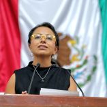 Diputada local con lentes amarillos habla desde la tribuna legislativa, frente a una bandera de México durante una sesión pública.