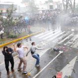 Jóvenes arrojan piedras hacia una línea de policías antimotines bajo chorros de agua a presión durante los disturbios en Guadalajara.