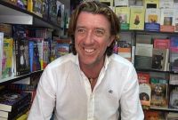 Colate Vallejo-Nágera sonríe durante su visita a la Feria del Libro, posando ante un expositor repleto de libros en un entorno cultural y literario.