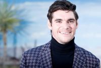 RJ Mitte, actor de Breaking Bad, sonríe con saco azul estampado y suéter negro de cuello alto frente a un fondo azul.