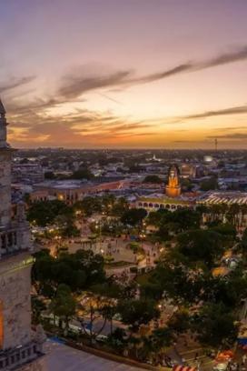 Panorámica aérea del centro histórico de Mérida, Yucatán, al atardecer, con la catedral y edificios coloniales iluminados, mostrando el entorno urbano y la actividad de la ciudad.