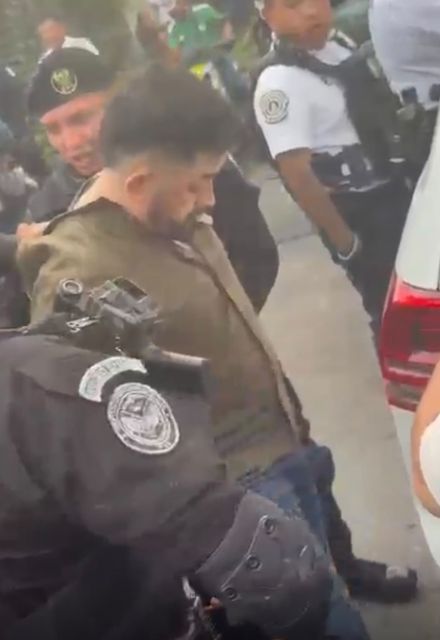 Hombre es sujetado por policías durante una detención junto a una camioneta, mientras otros elementos permanecen en el lugar.